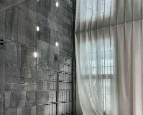 Conception lumière Smart Center à Tournai : mur en béton brut ponctué d'encastrés et voilage blanc éclairé en lumière naturelle