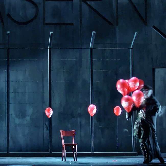 Scène de Wozzeck devant un mur de caserne gris-bleu et une clôture barbelée, ballons rouges et chaise rouge, Opéra de Nantes, conception lumière Ponctuelle